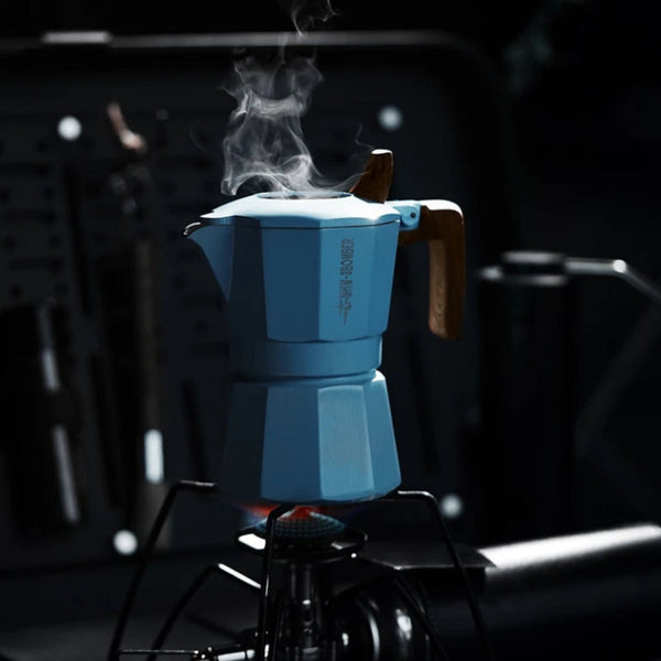 A light blue moka pot with a wooden handle sits on a portable stove, with steam rising from the pot against a dark, moody background.