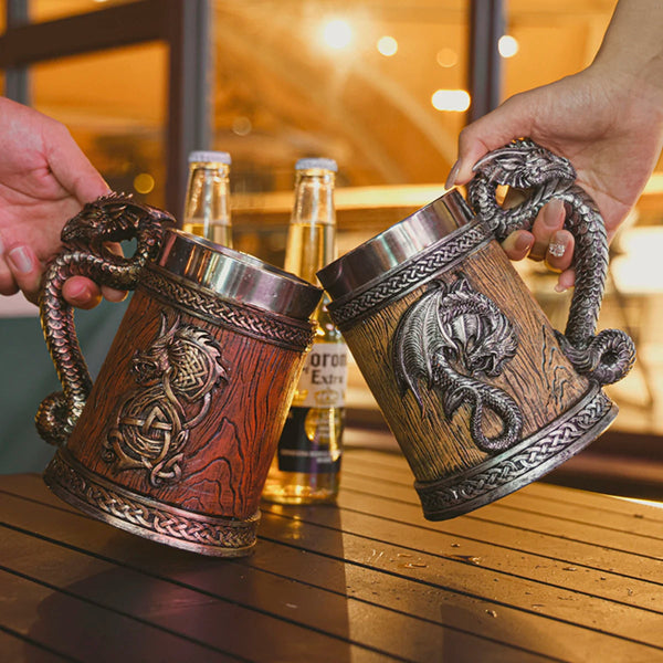 Two people clinking medieval dragon barrel tankards together in a cheers, showcasing the classic bronze and antique silver color variants at an outdoor bar setting with blurred beer bottles.