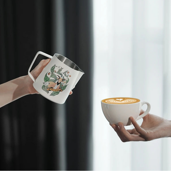 Hand holding an MHW-3BOMBER Origin Series milk pitcher next to a professional latte in a minimalist home setting.