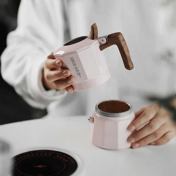 A person in a white long-sleeve top holds a light pink moka pot above another pink moka pot filled with coffee grounds, on a white tabletop.
