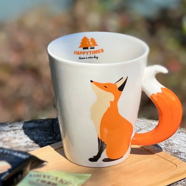 White ceramic coffee mug featuring an elegant standing fox illustration and a sculpted orange 3D fox tail handle on a wooden tray.