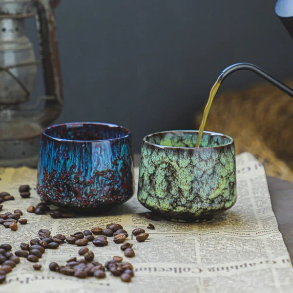 Pouring coffee into a minimalist green kiln-glazed ceramic cup next to a plum red espresso mug on newspaper with coffee beans.