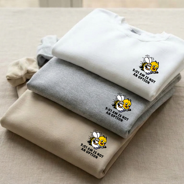 Three folded sweatshirts stacked on a beige fabric surface: a ceramic white sweatshirt on top with a small bee graphic and the text '9:01 IS NOT AN OPTION,' a medium heather ash grey sweatshirt in the middle, and an oatmeal cream sweatshirt on the bottom. A matching oatmeal cream sweatshirt is partially visible on the left.
