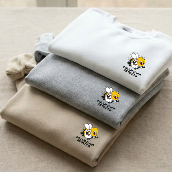 An Oatmeal Cream, Heather Ash Grey, Ceramic White crewneck sweatshirts with a small graphic on the left chest showing a premium corporate bee and the slogan 9:01 AM Is Not An Option'' display in a stack.