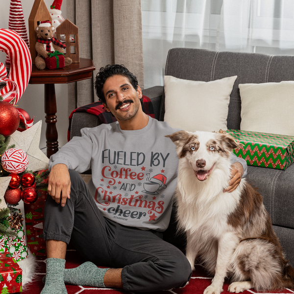 A man with a mustache sitting on a red geometric rug next to a Christmas tree and a Border Collie dog, wearing a grey crewneck sweatshirt that reads "Fueled by Coffee and Christmas Cheer" with festive red lettering and coffee cup graphics.