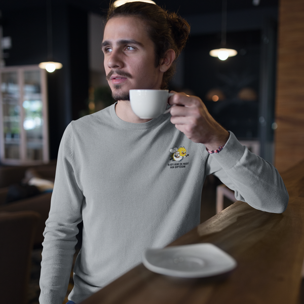 A young man with a bun and beard stands leaning against a dark wooden bar in a modern coffee shop, holding a white ceramic coffee cup to his lips. He is wearing the Heather Ash Grey crewneck sweatshirt from The Daily Buzz Series, featuring the premium black and yellow corporate bee emblem embroidered on the left chest with the text slogan 9:01 AM IS NOT AN OPTION.