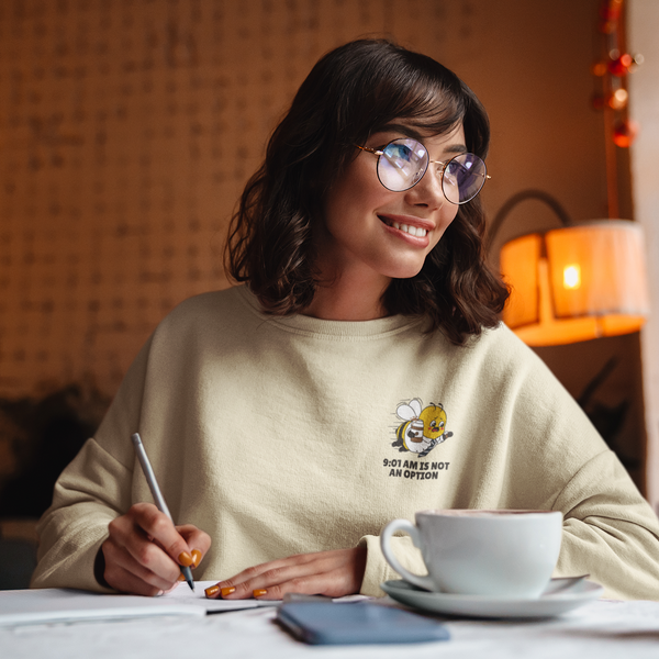 A young woman with round glasses smiles while writing in a notebook at a warm, cozy cafe table next to a white ceramic coffee mug. She is wearing the Sand Oatmeal Cream crewneck sweatshirt from The Daily Buzz Series, showcasing the premium black and yellow corporate bee emblem embroidered on the left chest with the text slogan 9:01 AM IS NOT AN OPTION.