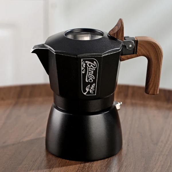 A black stovetop moka pot with a wooden handle and a silver 'Prince' logo on the side, sitting on a wooden table.